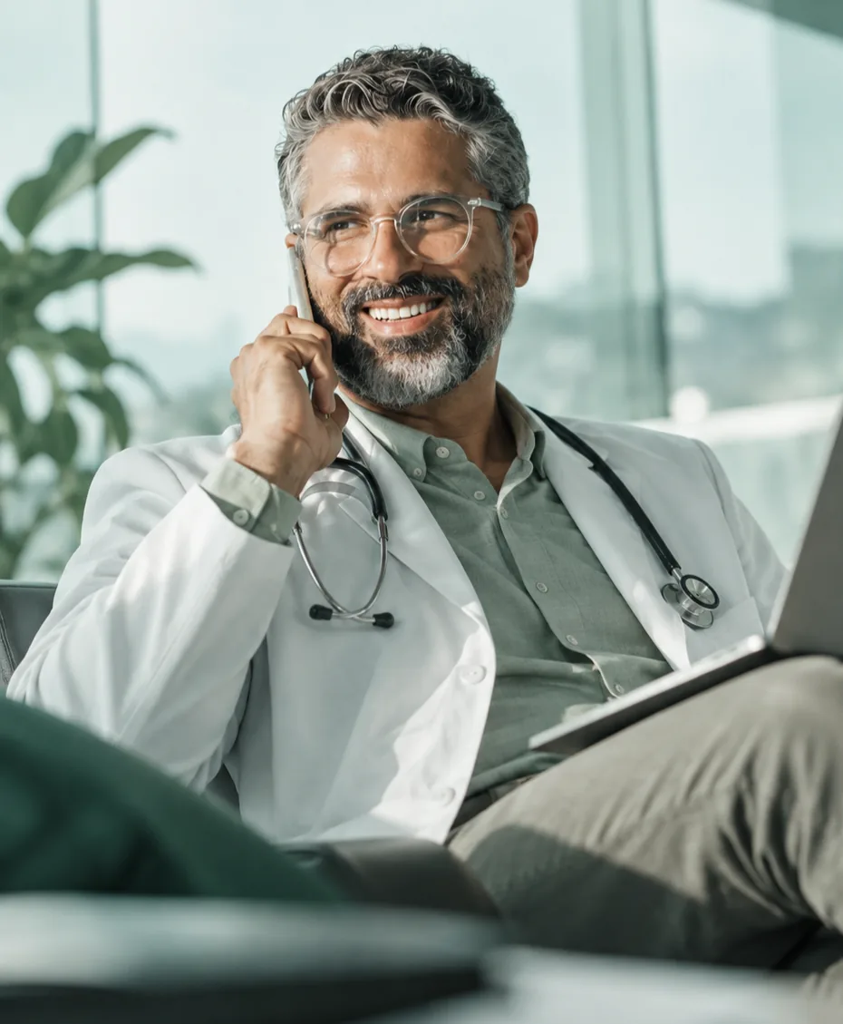 Médico de jaleco e estetoscópio sorrindo ao telefone, com um tablet no colo